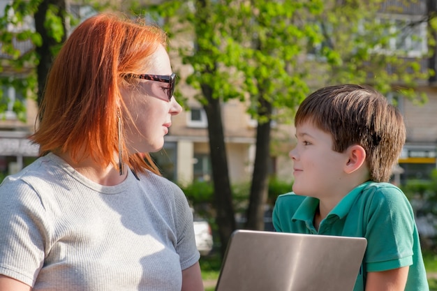 Jovem mulher ruiva no parque com seu filho pequeno no parque. Mãe e filho se olham. Mulher com laptop tentando trabalhar no parque e combinar trabalho e caminhada com uma criança