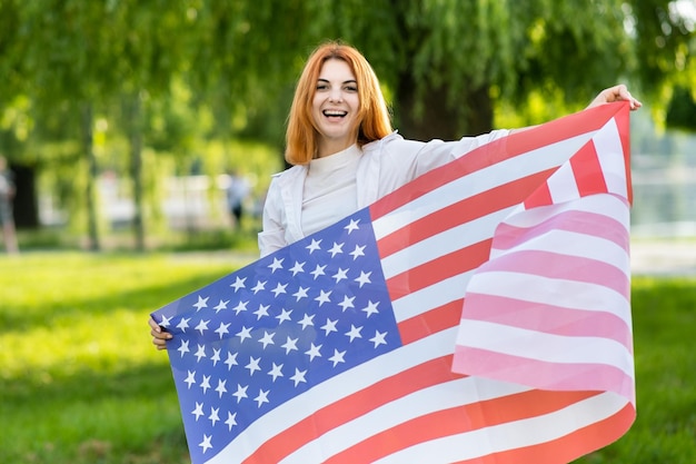 Jovem mulher ruiva segurando a bandeira nacional dos eua em pé ao ar livre no parque de verão. menina positiva comemorando o dia da independência dos estados unidos. dia internacional do conceito de democracia.