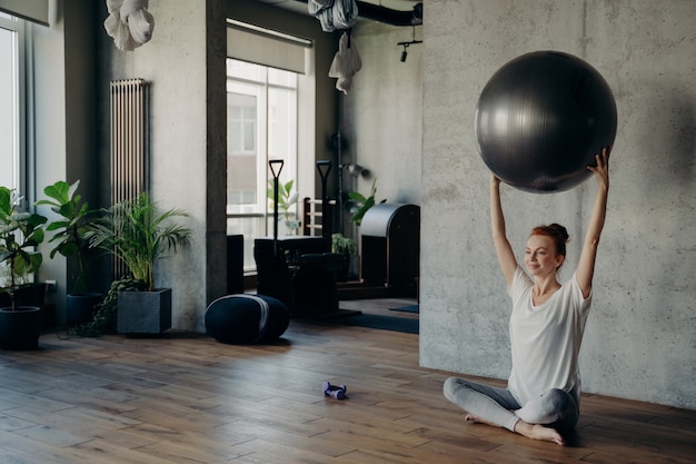 Jovem mulher ruiva sorridente, fazendo exercícios no estúdio de fitness, segurando uma grande bola de prata para exercícios com as duas mãos acima da cabeça e desfrutando de um estilo de vida saudável enquanto está sentado em posição de lótus no chão