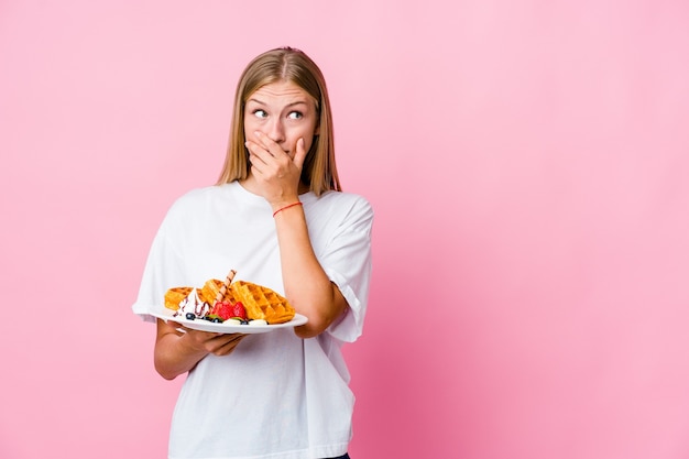 Jovem mulher russa comendo um waffle isolado pensativo, olhando para um espaço de cópia, cobrindo a boca com a mão.