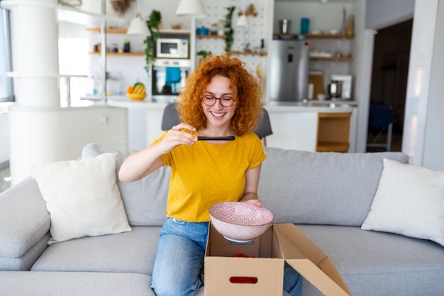 Jovem mulher satisfeita, feliz, viciada em compras, cliente sentada no sofá, desembalando a caixa de entrega de encomendas, conceito de envio de compras on-line, tirando fotos do produto para postar nas mídias sociais