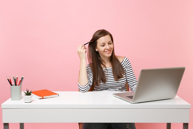 Jovem mulher satisfeita segurando um lápis trabalhando no projeto enquanto está sentado no escritório com o laptop pc isolado em fundo rosa pastel. Conceito de carreira empresarial de realização. Copie o espaço para anúncio.