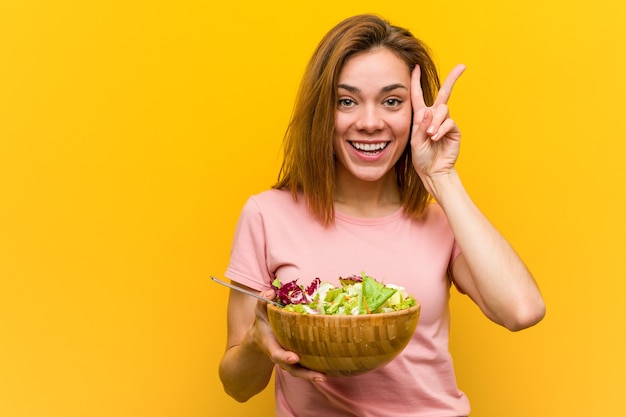 Foto jovem mulher saudável segurando uma salada, mostrando o sinal da vitória e sorrindo amplamente.