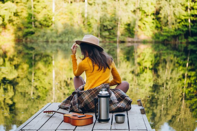 Foto jovem mulher se senta com uma guitarra em uma ponte em um lago com uma paisagem de outono. toning.