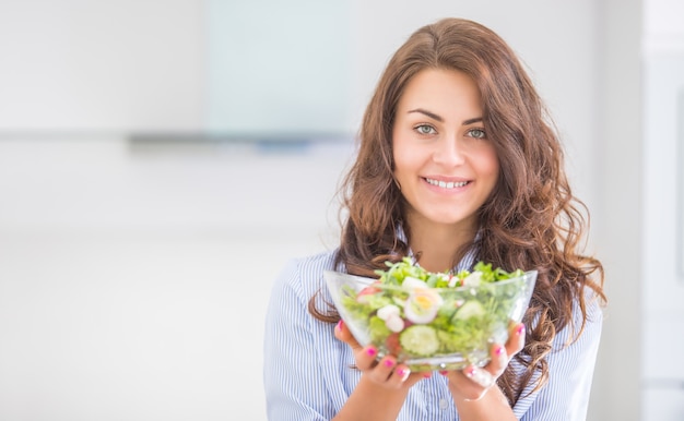 Jovem mulher segurando a tigela com salada na cozinha dela. Mulher bonita do conceito de estilo de vida saudável com vegetais misturados.