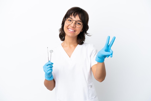 Jovem mulher segurando ferramentas isoladas no fundo branco, sorrindo e mostrando o sinal da vitória