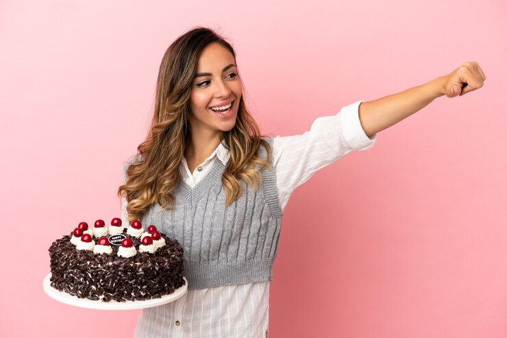 Foto jovem mulher segurando um bolo de aniversário sobre um fundo rosa isolado e fazendo um gesto de polegar para cima