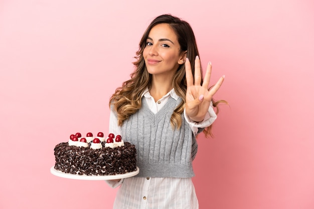 Jovem mulher segurando um bolo de aniversário sobre um fundo rosa isolado feliz e contando quatro com os dedos