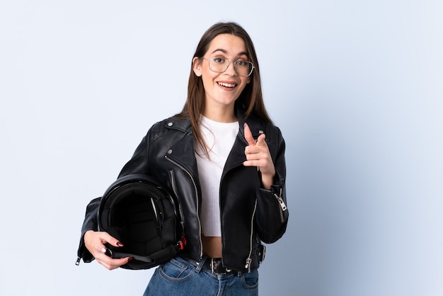 Jovem mulher segurando um capacete de moto sobre parede azul isolada aponta o dedo para você