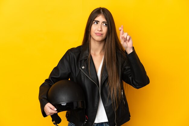 Jovem mulher segurando um capacete de motociclista isolado na parede amarela com os dedos se cruzando e desejando o melhor