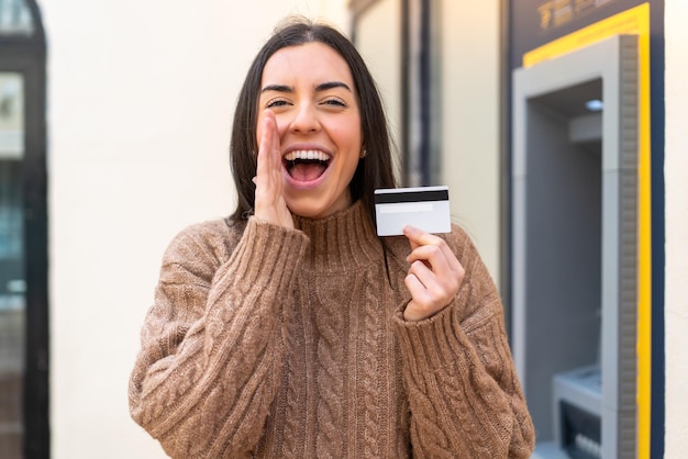 Jovem mulher segurando um cartão de crédito ao ar livre gritando com a boca aberta