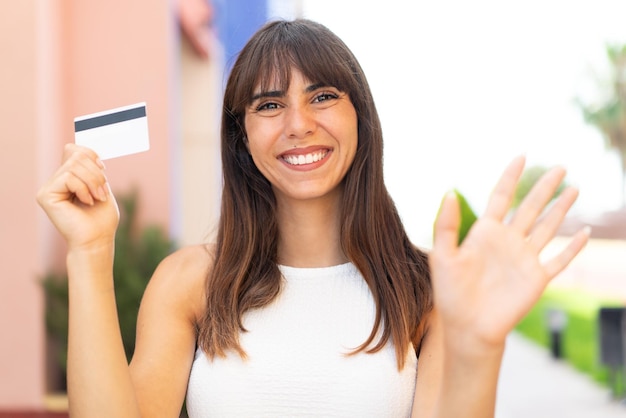 Jovem mulher segurando um cartão de crédito ao ar livre saudando com a mão com expressão feliz