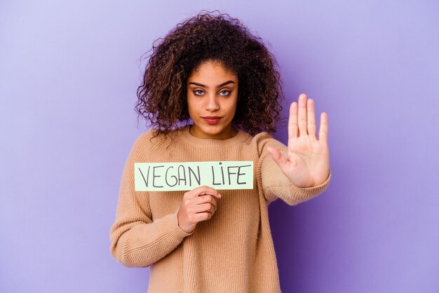 Jovem mulher segurando um cartaz da vida vegana isolada em pé com a mão estendida mostrando a placa de pare, impedindo você