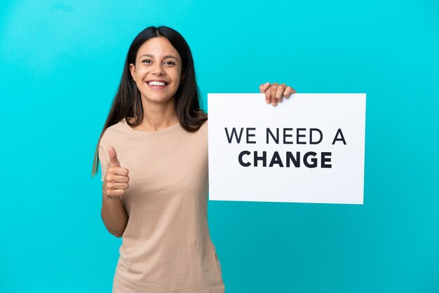 Jovem mulher segurando um fundo isolado segurando um cartaz com o texto Precisamos de uma mudança com o polegar para cima