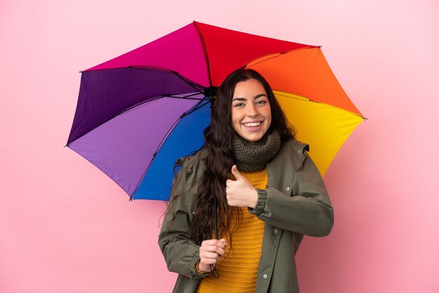 Jovem mulher segurando um guarda-chuva isolado em um fundo rosa e fazendo um gesto de polegar para cima