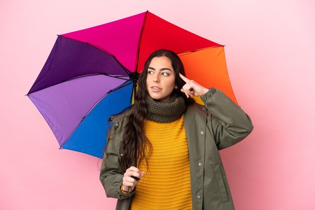 Jovem mulher segurando um guarda-chuva isolado em um fundo rosa, tendo dúvidas e pensando