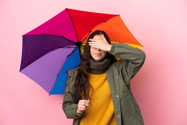 Jovem mulher segurando um guarda-chuva isolado no fundo rosa, cobrindo os olhos com as mãos. não quero ver nada