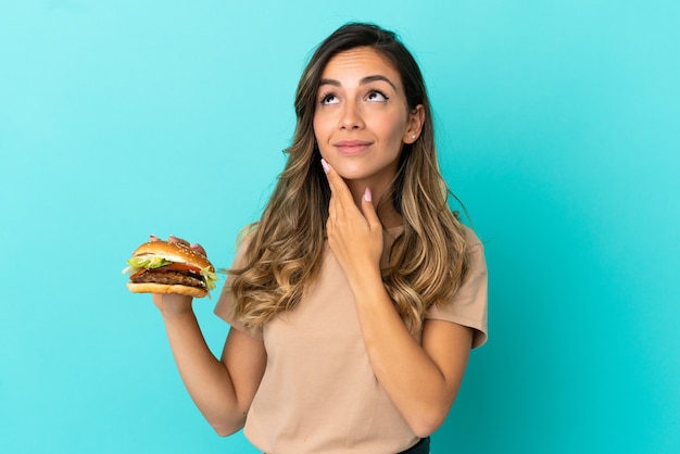 Jovem mulher segurando um hambúrguer sobre um fundo isolado, olhando para cima enquanto sorri