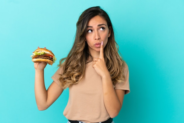 Jovem mulher segurando um hambúrguer sobre um fundo isolado, tendo dúvidas enquanto olha para cima