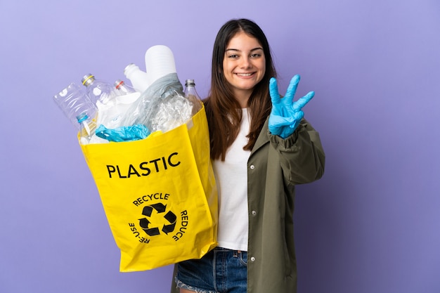 Jovem mulher segurando um saco cheio de garrafas de plástico para reciclar isolado no roxo feliz e contando três com os dedos