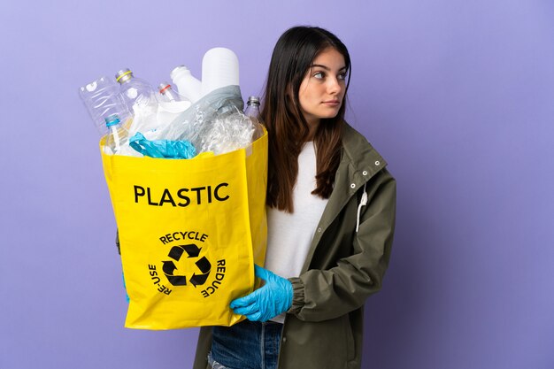 Jovem mulher segurando um saco cheio de garrafas de plástico para reciclar isolado no roxo, olhando para o lado