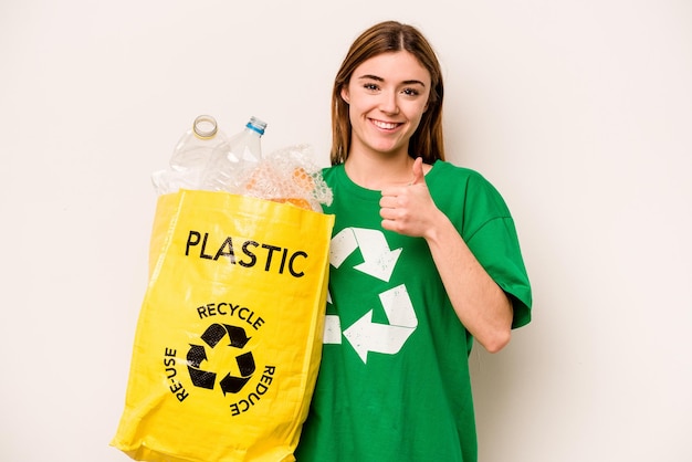 Jovem mulher segurando um saco cheio de garrafas plásticas para reciclar isolado no fundo branco sorrindo e levantando o polegar