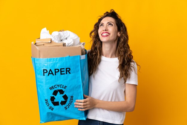 Jovem mulher segurando um saco de reciclagem cheio de papel para reciclar isolado em amarelo tendo uma ideia enquanto olha para cima