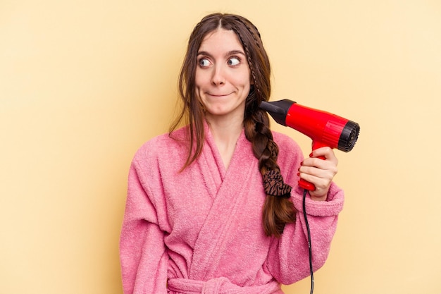 Jovem mulher segurando um secador de cabelo isolado em fundo amarelo confuso sente-se duvidoso e inseguro