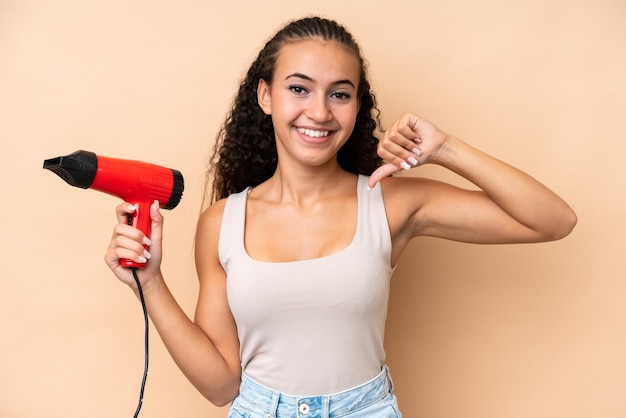 Jovem mulher segurando um secador de cabelo isolado em fundo bege orgulhoso e satisfeito