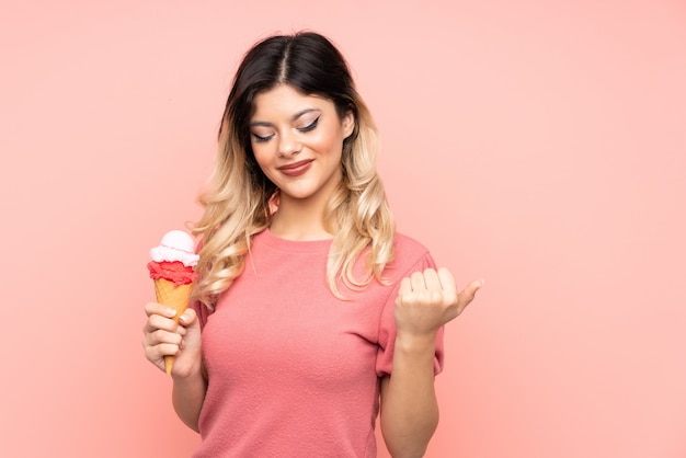 Jovem mulher segurando um sorvete de corneta isolado na parede rosa apontando para o lado para apresentar um produto