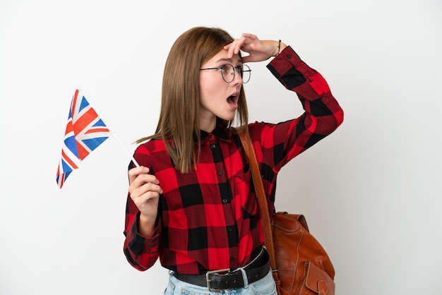 Jovem mulher segurando uma bandeira do Reino Unido isolada em fundo azul fazendo gesto de surpresa enquanto olha para o lado