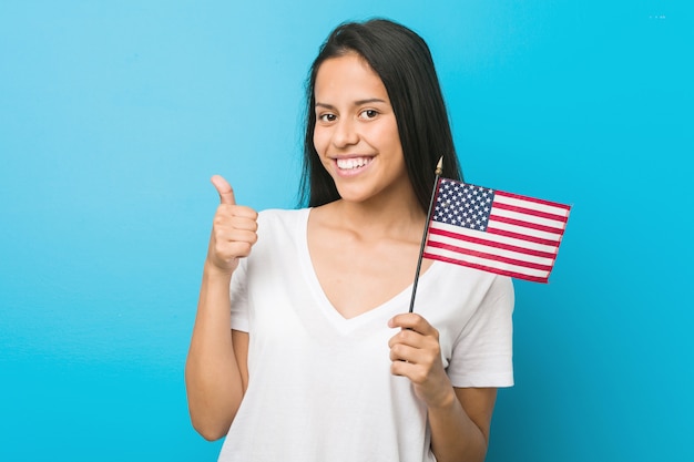 Jovem mulher segurando uma bandeira dos Estados Unidos, sorrindo e levantando o polegar