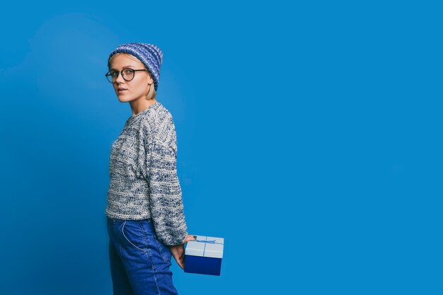 Jovem mulher segurando uma caixa de presente azul, escondida nas costas, vestida de azul contra uma parede azul.