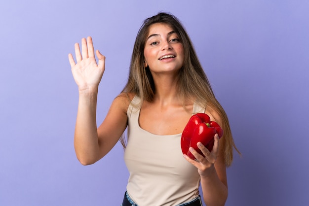 Jovem mulher segurando uma pimenta isolada em roxo saudando com mão com expressão feliz