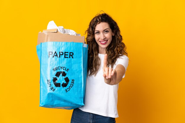 Jovem mulher segurando uma sacola cheia de papel para reciclar, isolada na parede amarela, mostrando e levantando um dedo