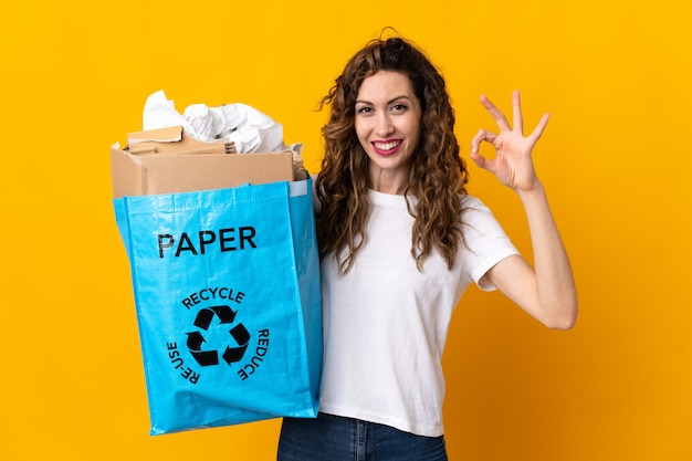 Jovem mulher segurando uma sacola cheia de papel para reciclar, isolada na parede amarela, mostrando um sinal de ok com os dedos
