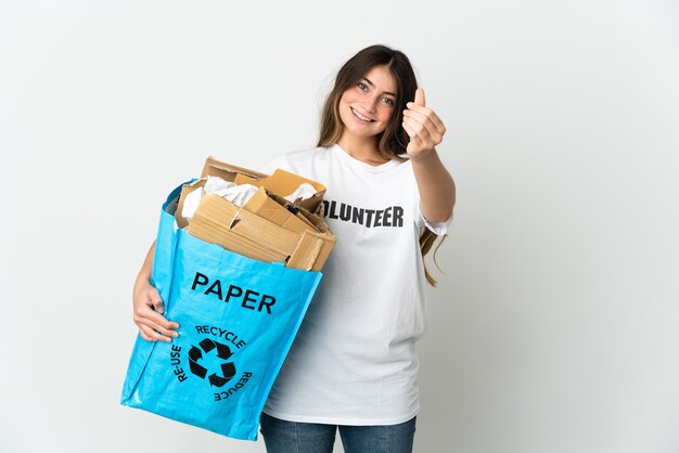 Jovem mulher segurando uma sacola cheia de papel para reciclar isolada na parede branca fazendo gesto de dinheiro