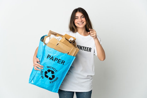 Jovem mulher segurando uma sacola cheia de papel para reciclar isolada no branco surpresa e apontando para a frente