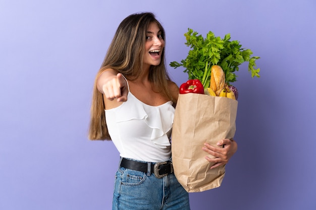 Jovem mulher segurando uma sacola de compras de supermercado apontando para a frente com uma expressão feliz