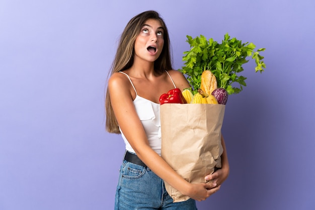 Jovem mulher segurando uma sacola de compras de supermercado, olhando para cima e com expressão de surpresa