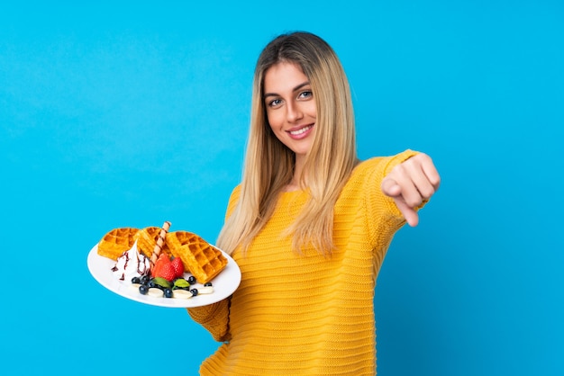 Jovem mulher segurando waffles aponta o dedo para você com uma expressão confiante