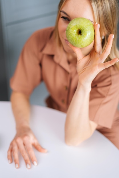 Jovem mulher sentada à mesa e segurando a maçã verde saborosa na frente dos olhos na cozinha com interior moderno. conceito de alimentação saudável.