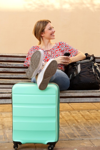 Jovem mulher sentada com bagagem sentada no banco com telefone móvel