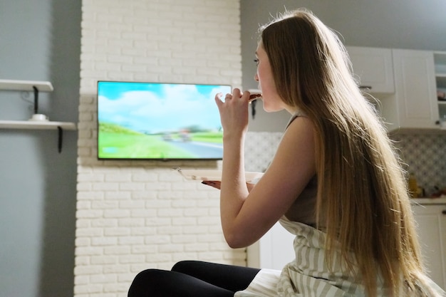 Jovem mulher sentada em casa assistindo tv e comendo