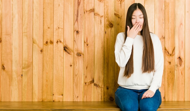 Jovem mulher sentada em um lugar de madeira bocejando mostrando um gesto cansado, cobrindo a boca com a mão