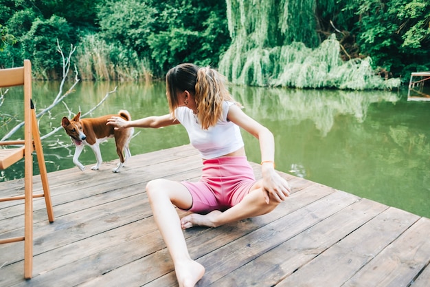 Jovem mulher sentada em um píer de madeira em um lago e brincando com seu cachorro.