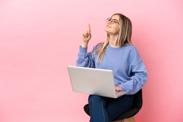 Jovem mulher sentada em uma cadeira com laptop sobre fundo rosa isolado apontando com o dedo indicador uma ótima ideia