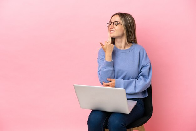 Jovem mulher sentada em uma cadeira com laptop sobre fundo rosa isolado apontando para o lado para apresentar um produto