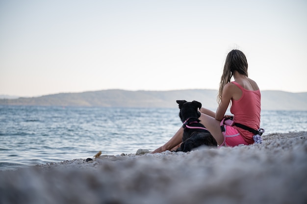 Jovem mulher sentada na bela praia de seixos com seu cão pastor preto deitado ao lado dela.