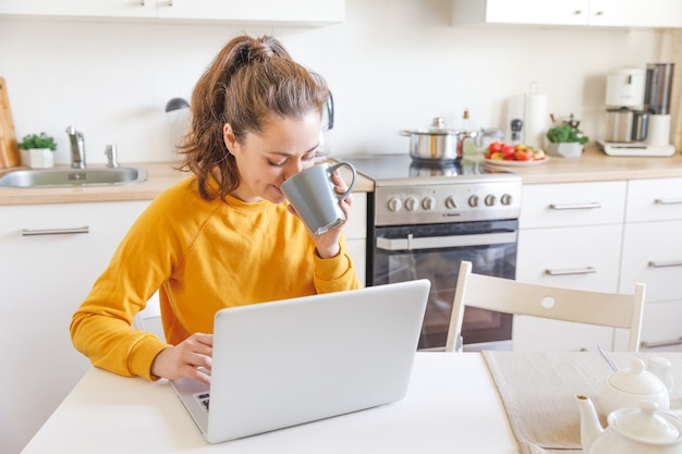 Jovem mulher sentada na cozinha em casa, trabalhando, usando um laptop